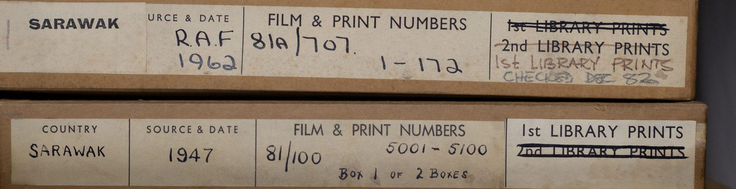 Six flat, tan coloured cardboard boxes are piled together. Each box has a label with printed and handwritten text on it. This text gives information, including locations dates and film and print numbers. The labels are slightly discoloured.