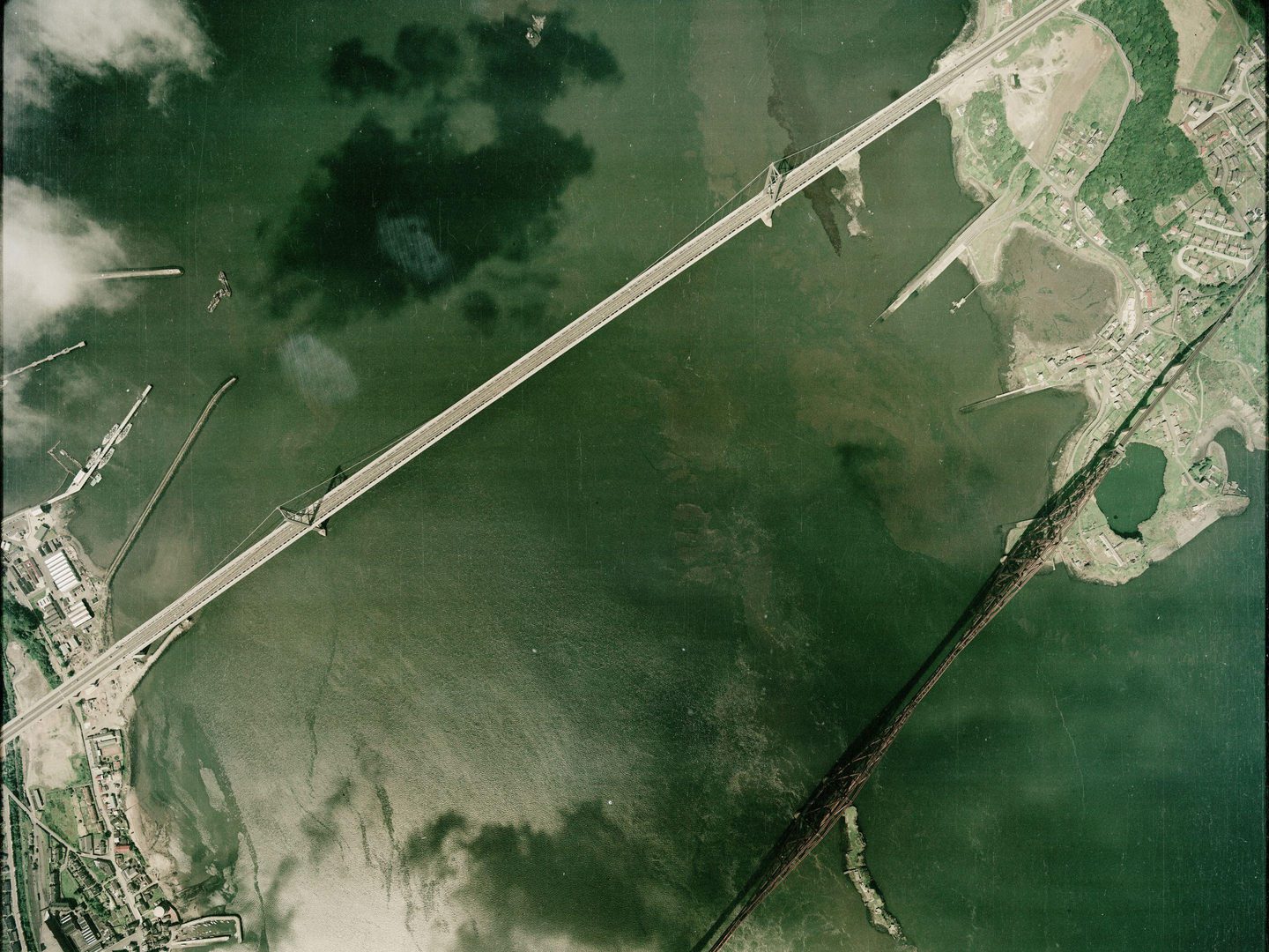 A colour aerial photo of the Forth bridges. Small areas of land are visible in the bottom left and top right corners, separated by a large area of greenish-looking sea. The areas of land are joined by a light grey road bridge and a red rail bridge.