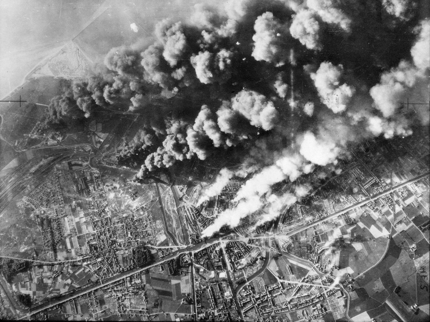 A black and white aerial photo of the city of Dunkirk, with the coast just visible in the distance. Thick clouds of black and white smoke are billowing up from the city, being driven by the wind from left to right.