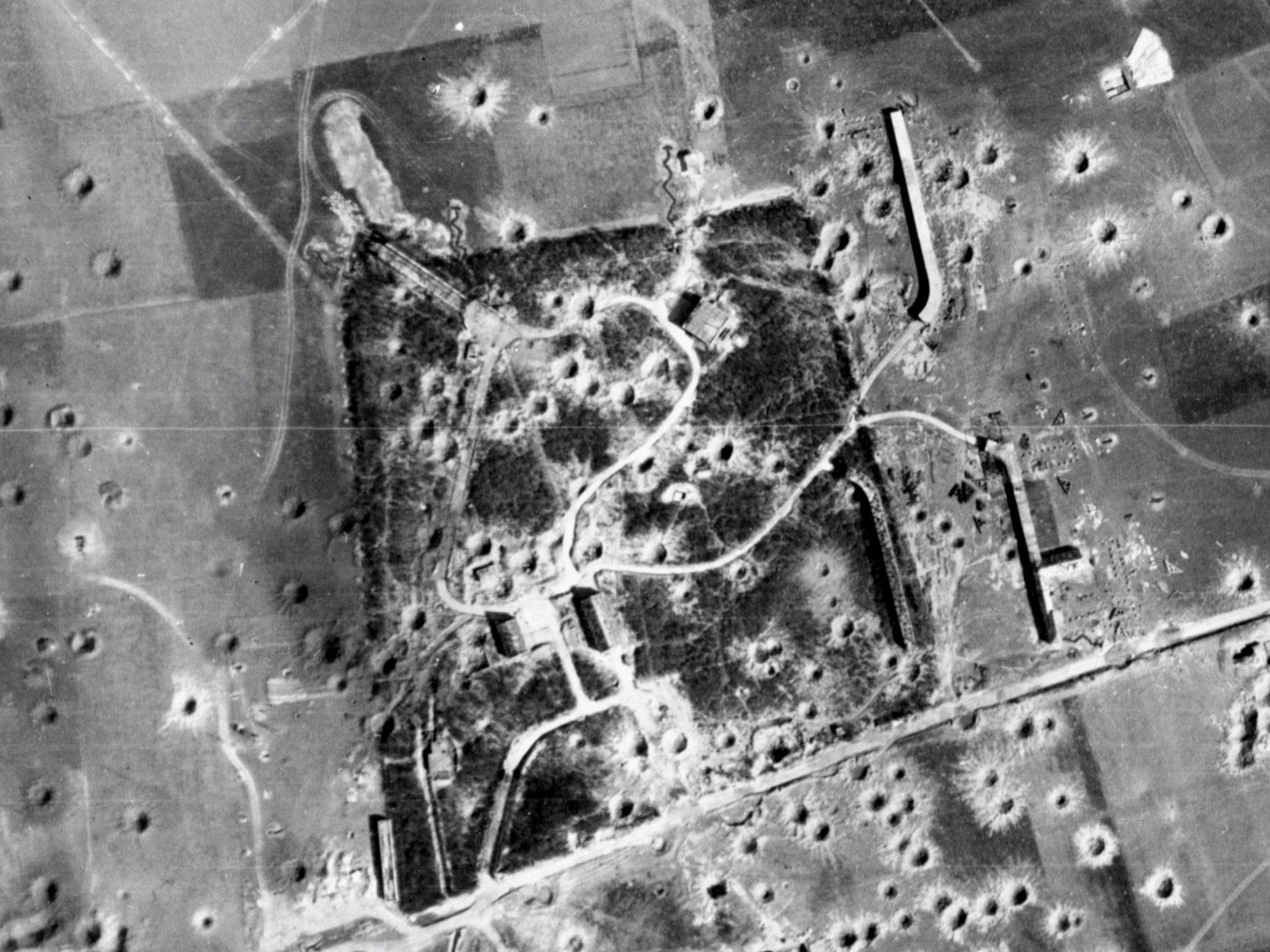 A black and white aerial photo of a 4-sided woodland surrounded by fields with a road running along the bottom edge. Buildings are visible within and to the right of the wood. The area has been bombed and craters are visible in the fields and wood.