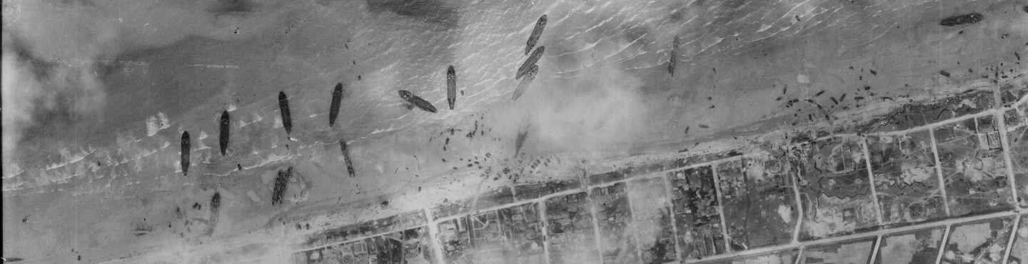 A black and white aerial photo of about a dozen ships pulled up on a beach. The land beyond the beach has been divided up into a grid, and the scene is slightly obscured by clouds or smoke.