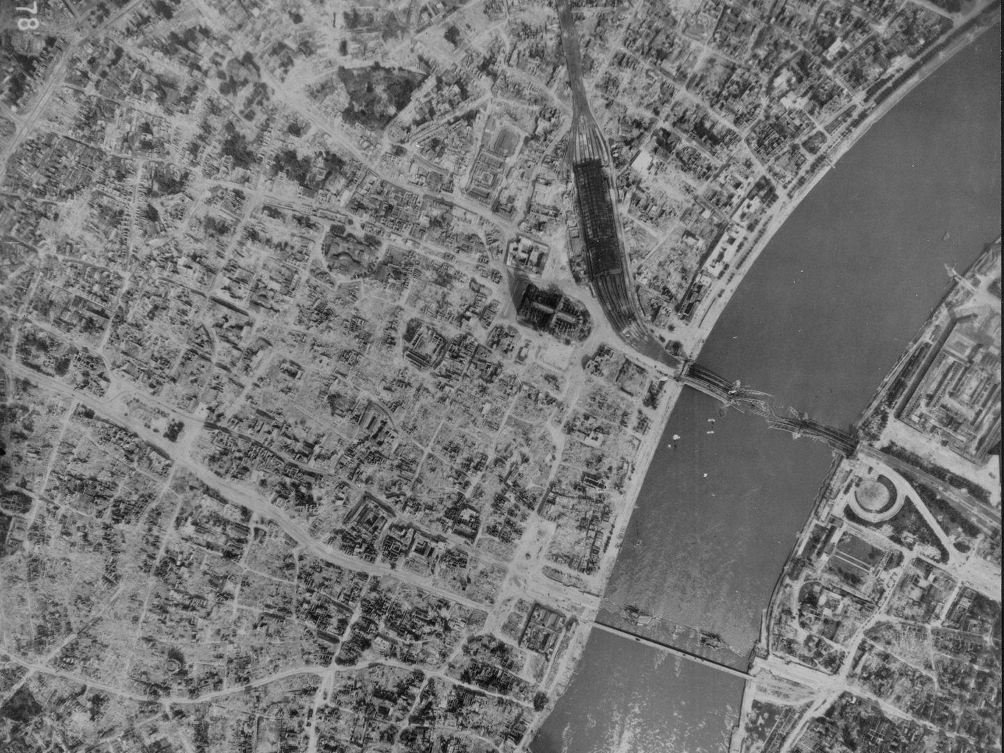 A black and white aerial photo of the bombed city of Cologne, with a wide river running along the right edge. The outlines of streets are visible but almost all the buildings have been destroyed. Cologne cathedral remains standing just above centre.