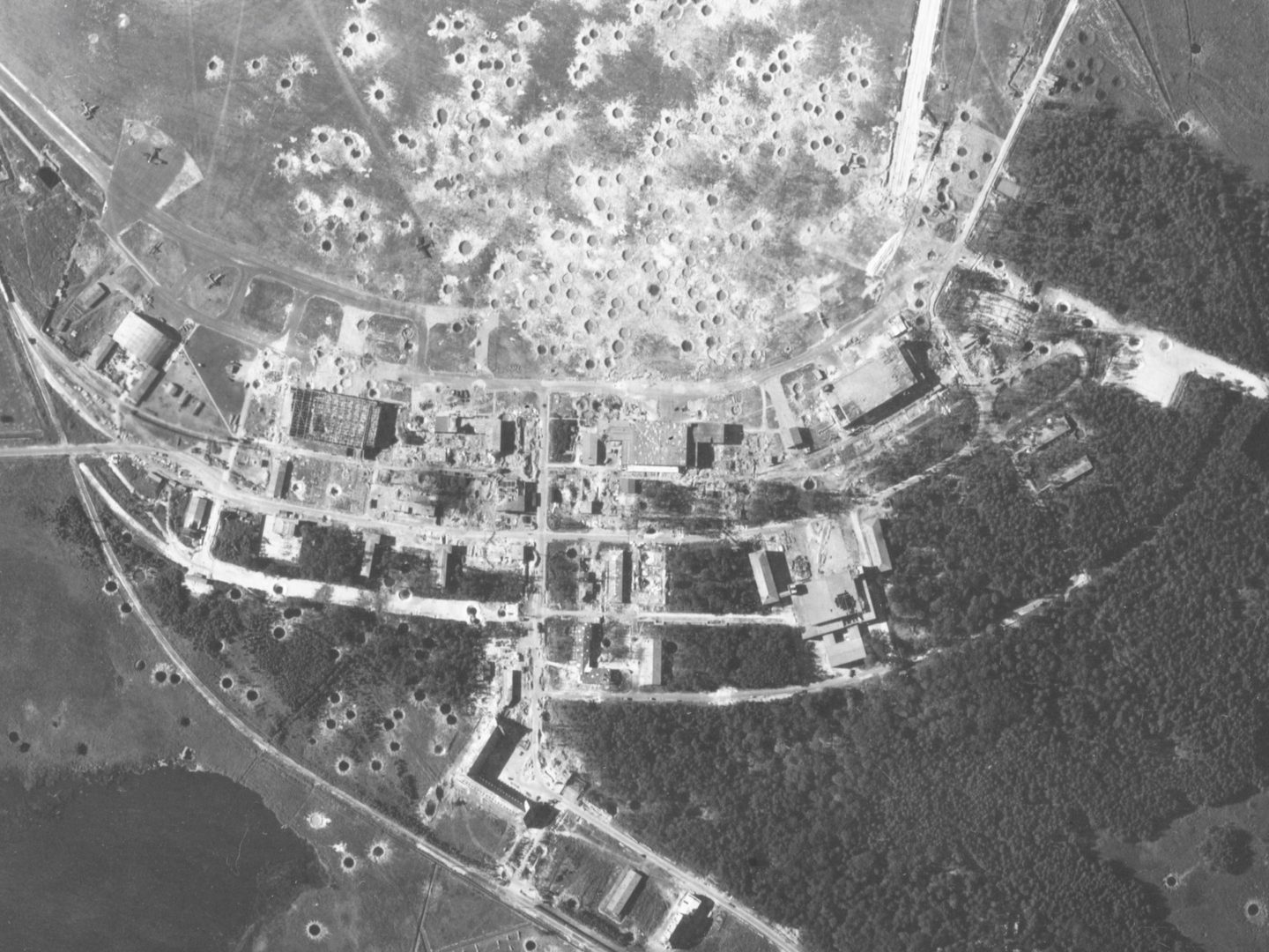 A black and white aerial photo of factory buildings amongst trees and fields. The area has been extensively bombed and craters are visible in the fields and throughout the buildings.