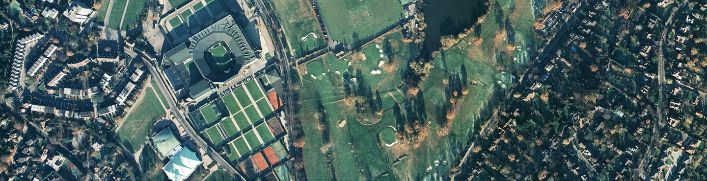 A colour aerial photo of the Wimbledon area in London. Streets of houses are visible at the right and left edges. A golf course with bunkers and trees occupies the middle. To the left of centre are about 20 tennis courts and a tennis stadium.