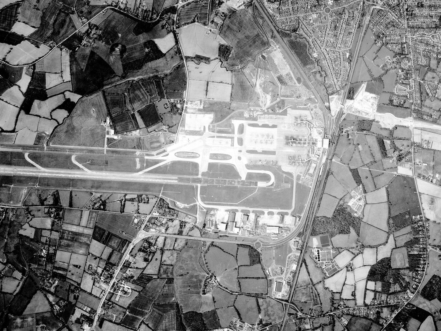 A black and white aerial photo of an airport surrounded by fields and some houses. The runway runs from the centre of the image to the left edge. Planes and airport buildings are visible. A motorway runs vertically through the image right of centre.