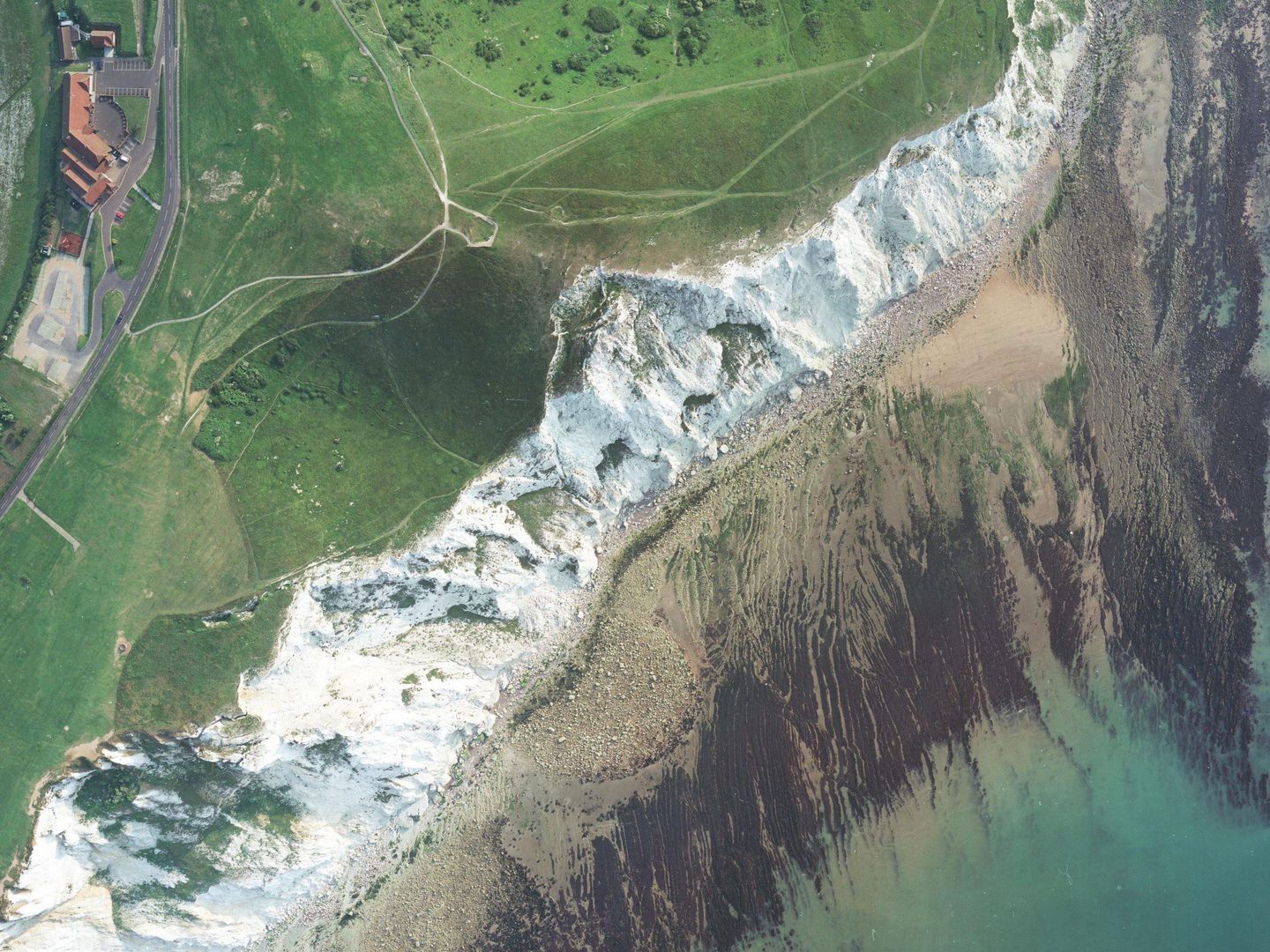 A colour aerial photo with white cliffs stretching from the bottom left corner to the top right. There is green grass at the top of the cliffs and a building in the top left corner. Below the cliffs is a rocky beach and a small area of turquoise sea.