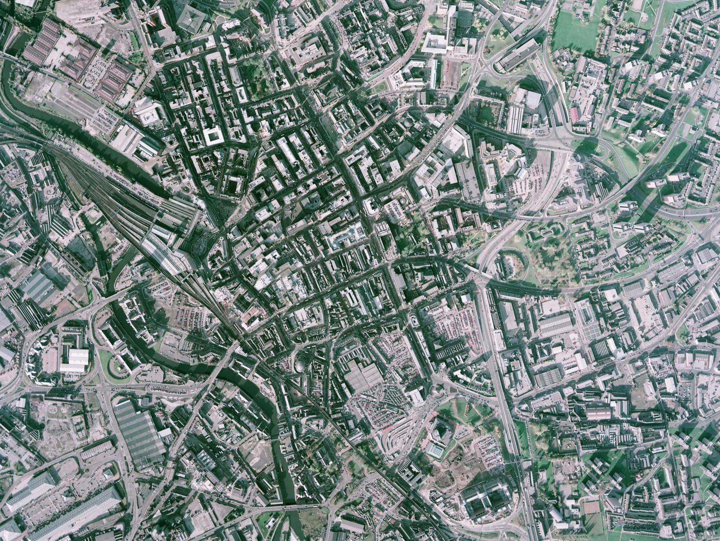 A slightly washed-out colour aerial photo of Leeds. The centre of the image shows large buildings with a train station to the left of centre. To the left are large structures which might be industrial buildings. To the right are several green spaces.