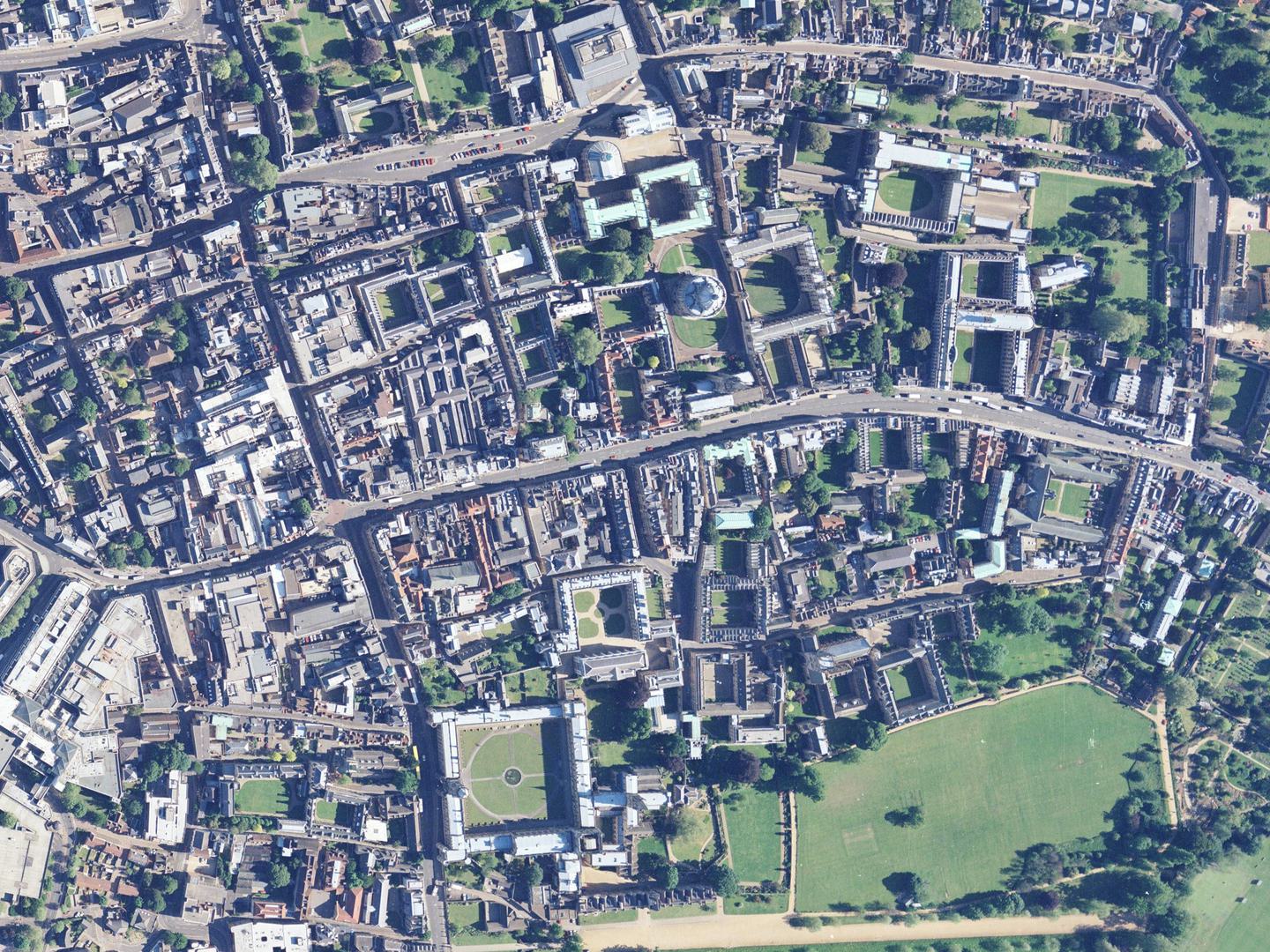 A colour aerial photo of Oxford, with buildings arranged around a road running from left to right. Spires and domes are visible, along with quadrangles of buildings arranged around grass. An open park surrounded by trees is at the bottom right.