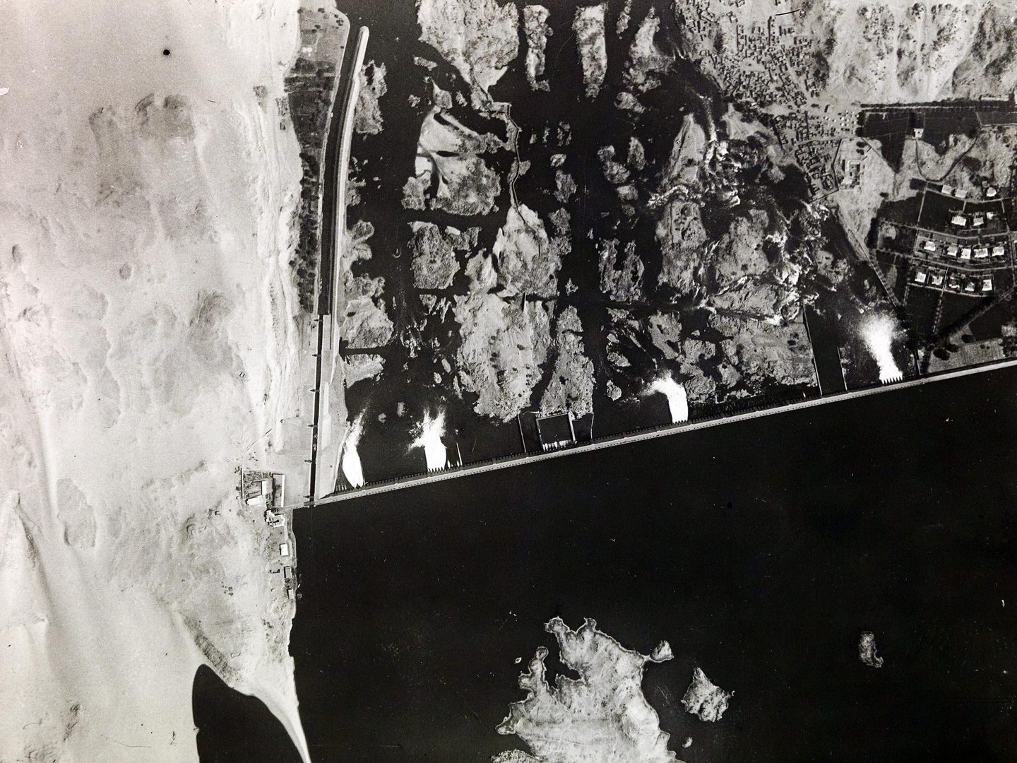 A black and white aerial photo of a dam in the desert. There is a large area of white sand along the left edge, and very black-looking water across the bottom third. Four torrents of water can be seen spilling onto the rocky ground below the dam.
