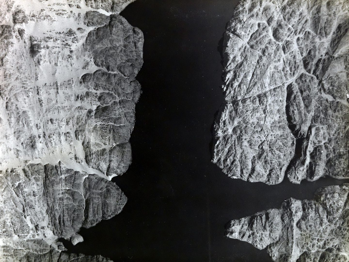 A black and white aerial photo of a lake. The lake runs from top to bottom through the middle of the image. The water looks very black. Either side are rocky shores, partially covered in sand. The shores are fissured with old water courses.