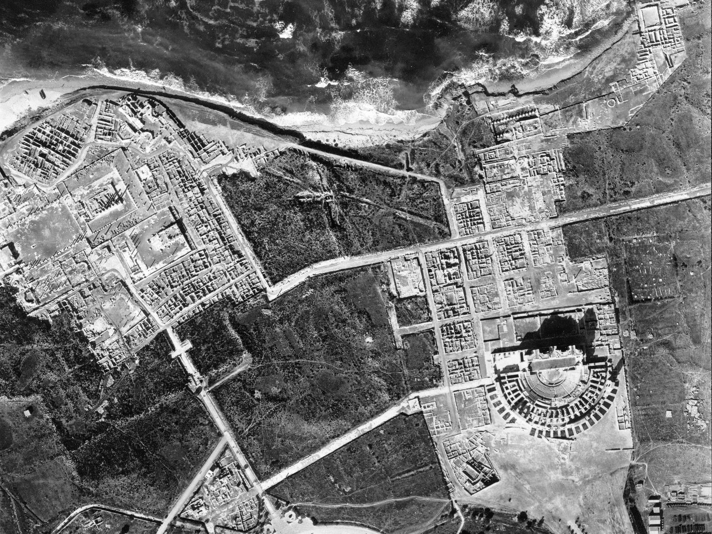 A black and white aerial photo of ancient ruins. The ruins are arranged in two distinct groups to the left and right. The right-hand group contains a semi-circular amphitheatre. The coast and a small strip of sea are visible at the top of the image.
