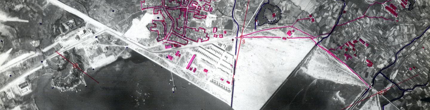 A black and white aerial photo of a city on the edge of the sea. There are buildings and an industrial area in the centre and fields to the right. The photo has been marked with purple pencil highlighting blocks of buildings.