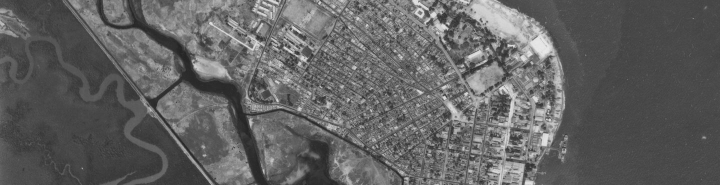 A black and white aerial photo of a city. The streets are arranged in a grid. There is a coastline to the right with an area of dark sea at the right-hand edge. To the left are two meandering watercourses and an area of undeveloped wetland.