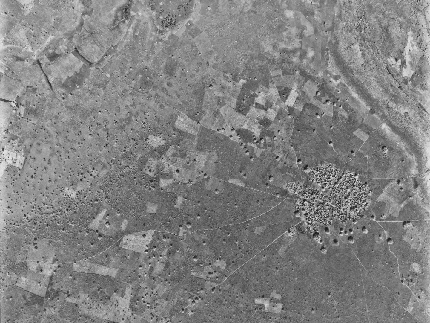 A black and white aerial photo of a village. The village is in the bottom right quadrant of the image and circular in shape. Paths radiate from the village into the tree-studded savannah surrounding it. Some fields are visible around the village.