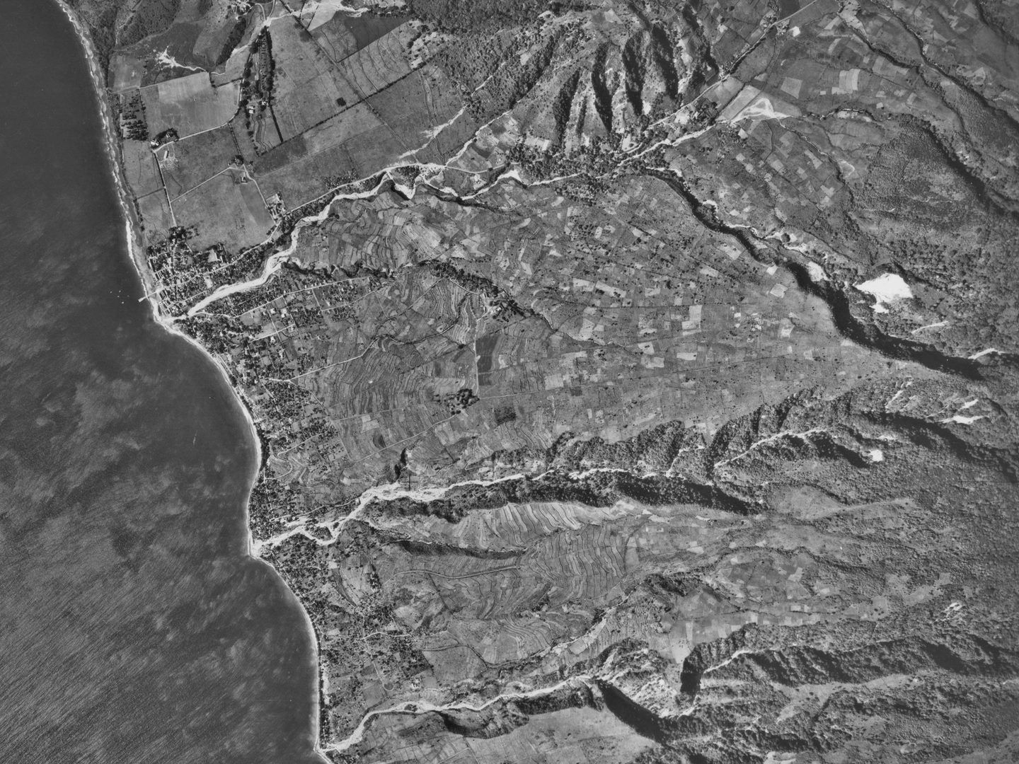 A black and white aerial photo of a coastline. The land to the right is mountainous, with three watercourses running down to the sea on the left. A scattered settlement is visible along the edge of the coast.