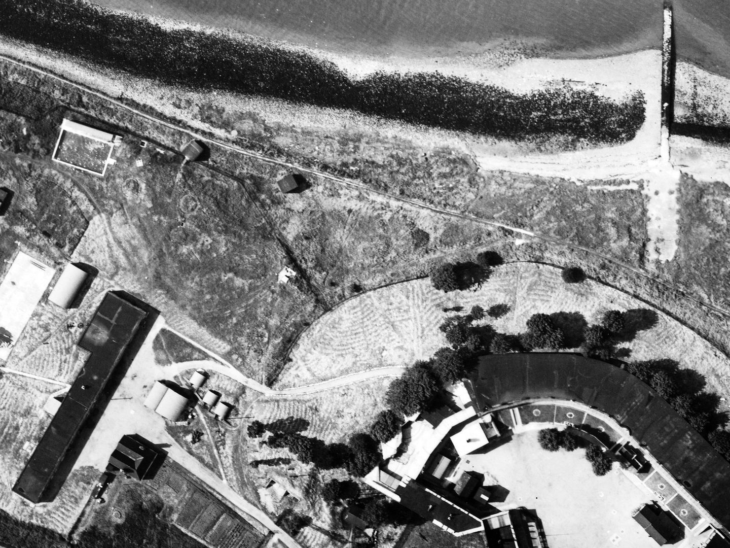 A black and white aerial photo of buildings on a coastline. The coast runs along the top edge, with a small amount of water visible right at the top. The buildings are arranged along the bottom edge and are interspersed with a few trees.