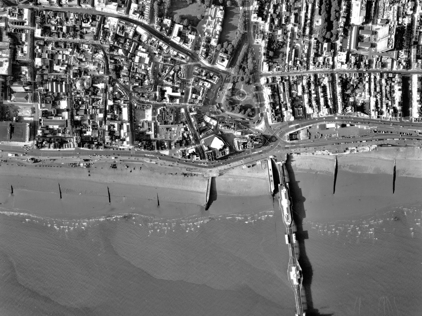 A black and white aerial photo with a city at the top half of the image and the sea at the bottom half. Right of centre a pier protrudes into the sea. Buildings and trees are visible on the land, with the domes of Brighton Pavilion at top middle.