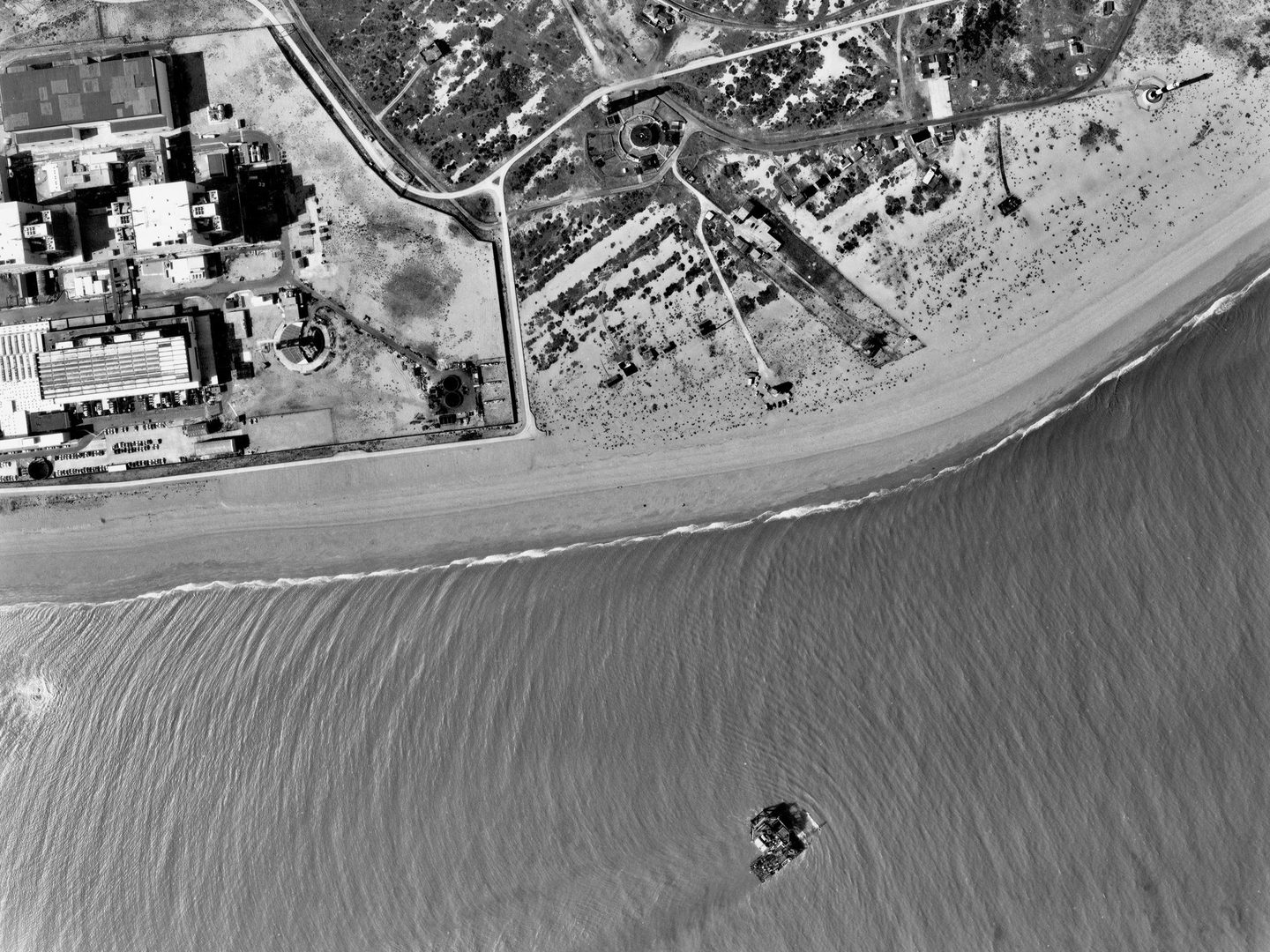 A black and white aerial photo of a coast, with land in the image’s top half and sea in the bottom half. The large buildings of Dungeness nuclear power station are visible to the left, and a pebble beach to the right. A structure stands in the sea.