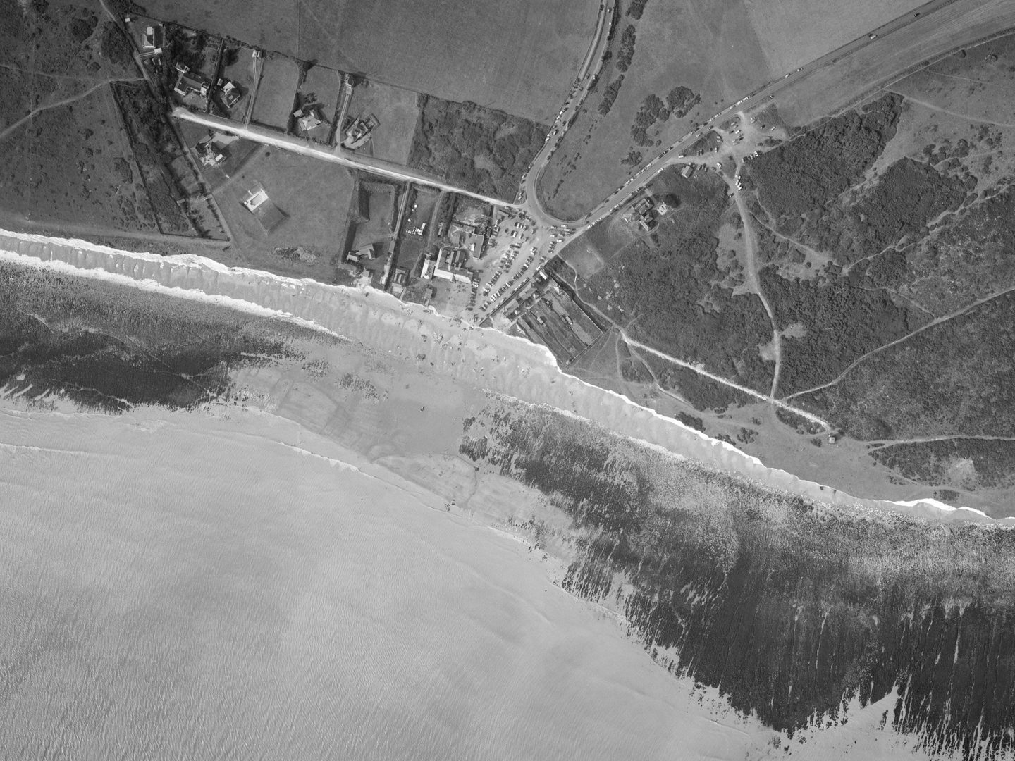 A black and white aerial photo of a coast, with land in the image’s top half and sea in the bottom half. Buildings, a carpark and a road are visible just above centre. The coastline looks rocky, and the sea is foaming white on the shore.