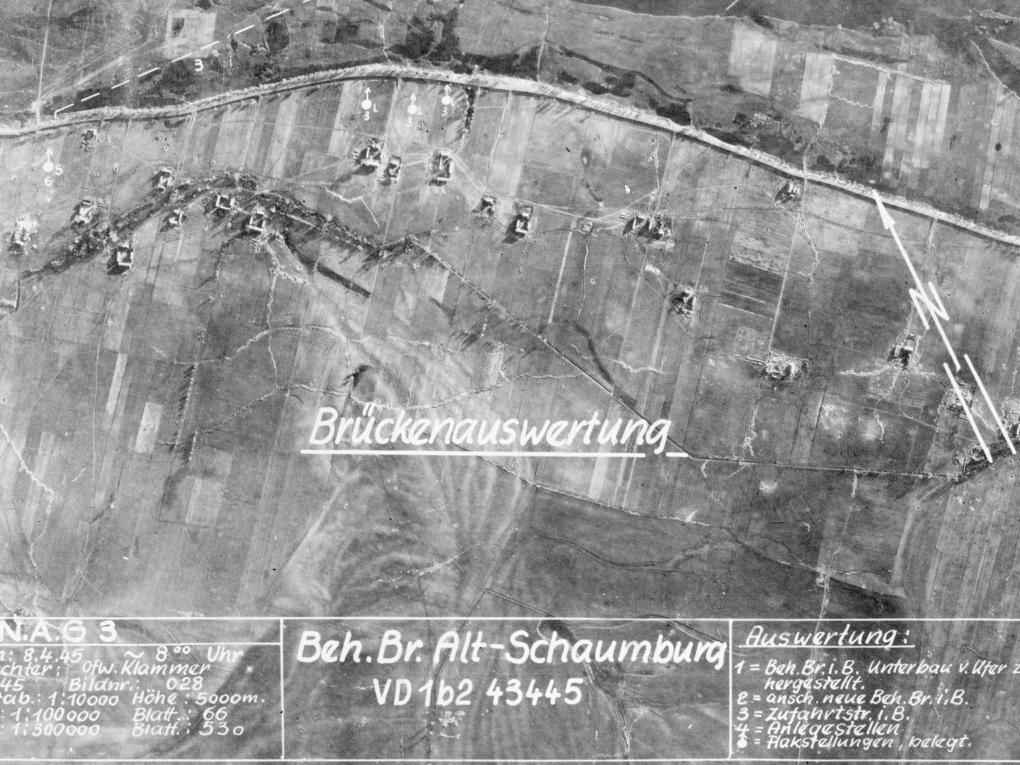 A black and white aerial photomap of fields. A road runs horizontally along the top edge, with ruined buildings alongside it. There are annotations across the image, German text along the bottom and the word ‘Brückenauswertung’ across the middle.