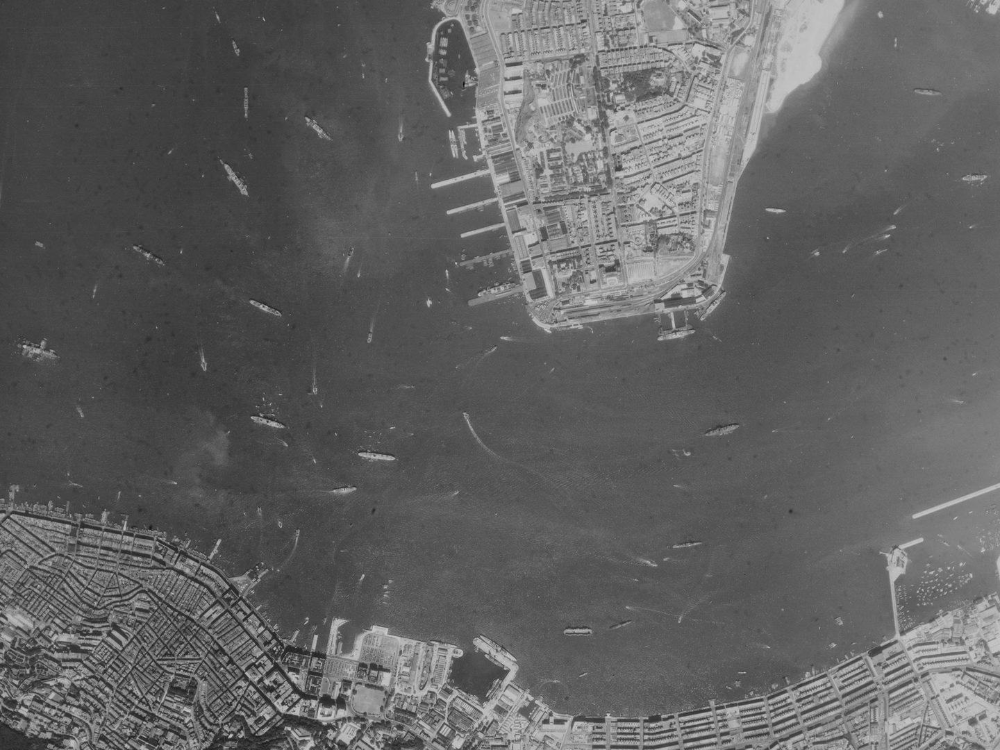 A black and white aerial photo of the sea, with numerous ships on it. A strip of heavily built-up land runs along the bottom edge. A peninsula of land protrudes into the sea from the top edge, with port buildings and piers protruding to the left.