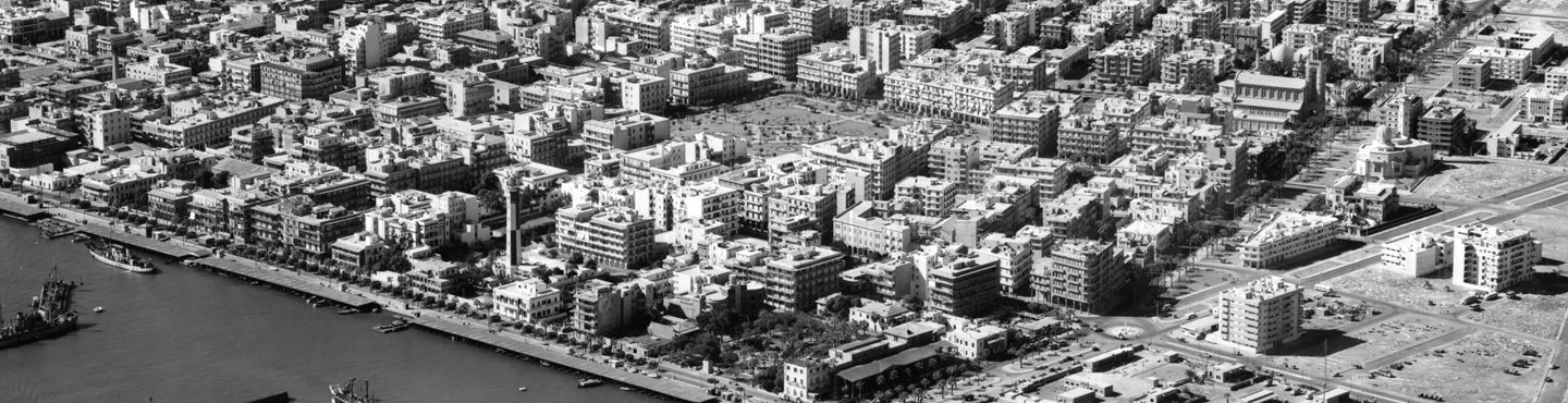 A black and white oblique aerial photo of a city with a coastline. The sea is at the bottom left corner. A small number of ships are on the water. The city is made up of what appears to be low-rise apartment blocks. There is a park in the centre.