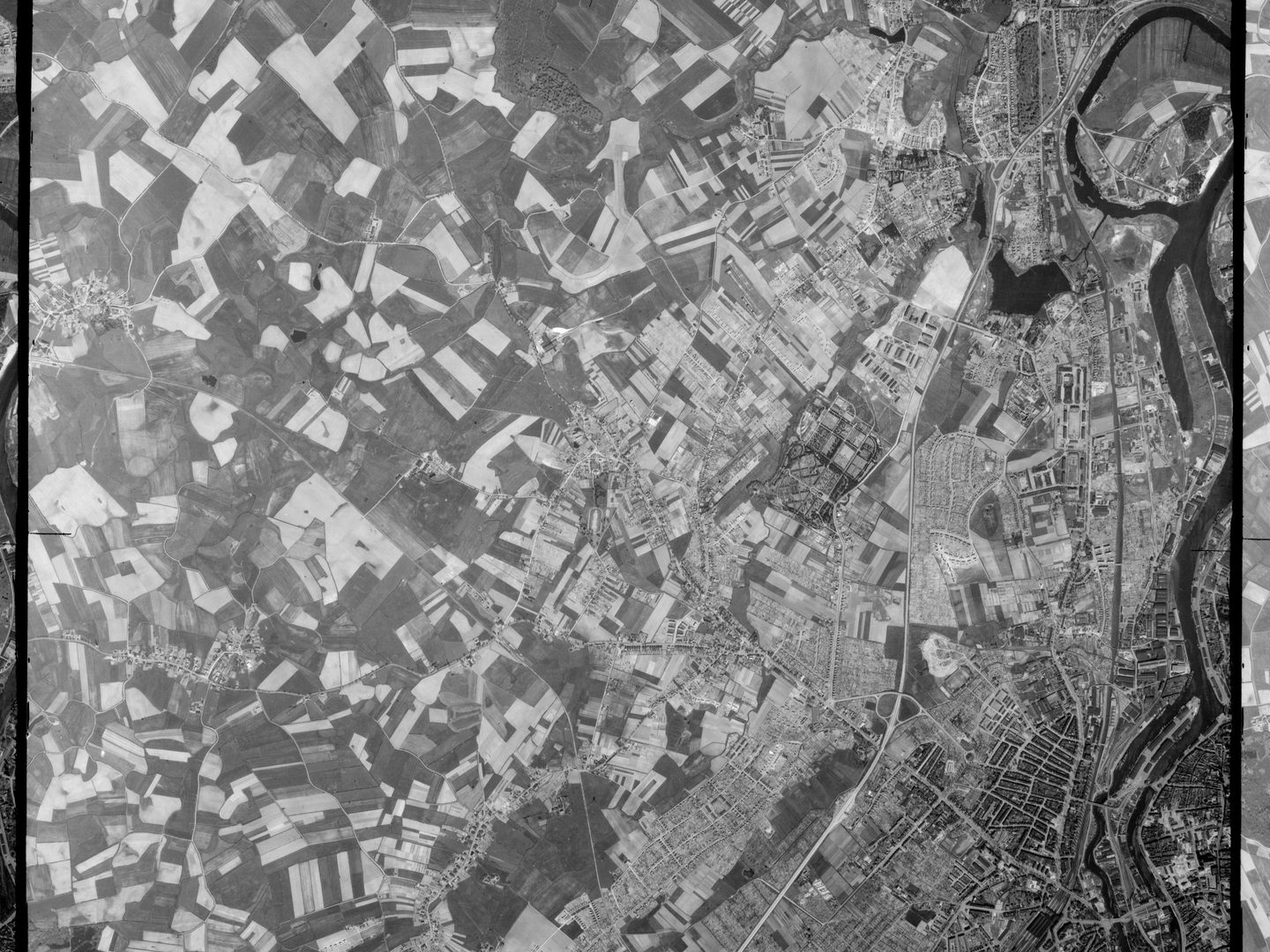A black and white aerial photo from high altitude, with an irregular grid of fields in light and dark colours across the left side, and the buildings of a town across the right side. A river runs from top to bottom along the very right-hand edge.