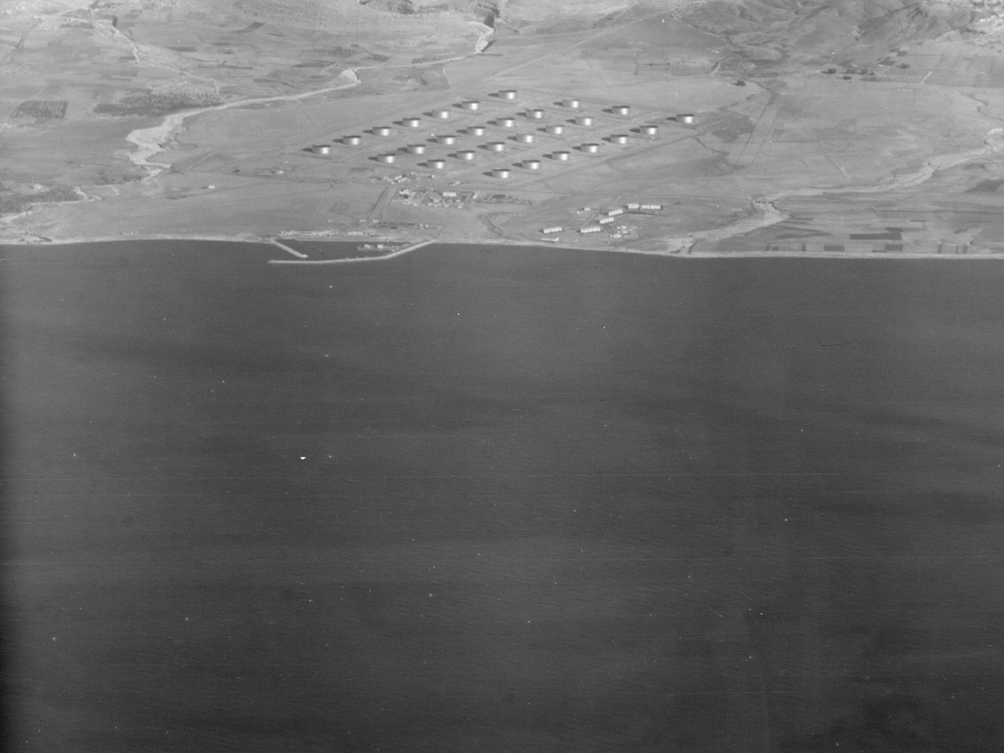 A black and white oblique aerial photo, with a large area of dark sea across the bottom two thirds. A pale-coloured shore is visible in the top third, with 28 shiny circular tanks arranged in a grid.