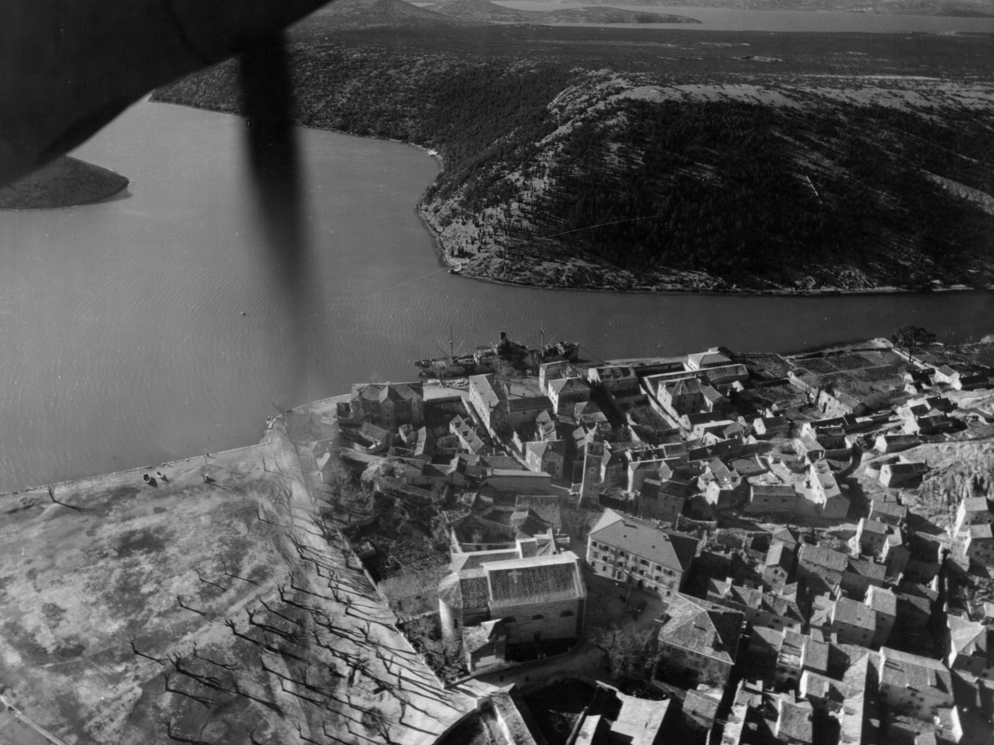 A black and white oblique aerial photo with scrub-covered hills in the background and water in the midground. The roofs of a town are visible in the foreground. In the top left corner a portion of plane engine and propeller is visible.