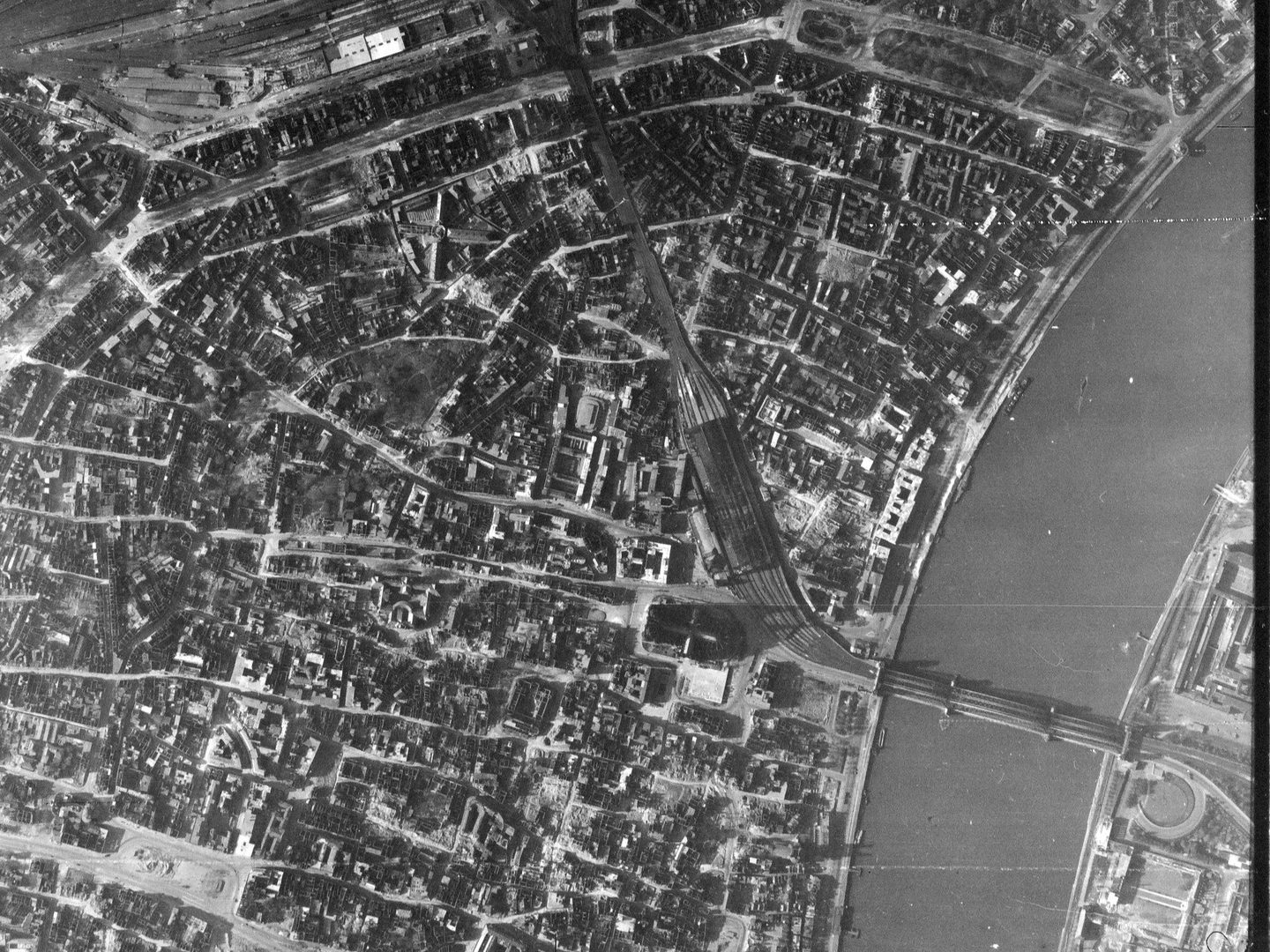 A black and white aerial photo of the city of Cologne, with a wide river crossed by a railway bridge on the right. The streets are visible, and some buildings have been damaged by bombing. Cologne cathedral is visible just below right of centre.