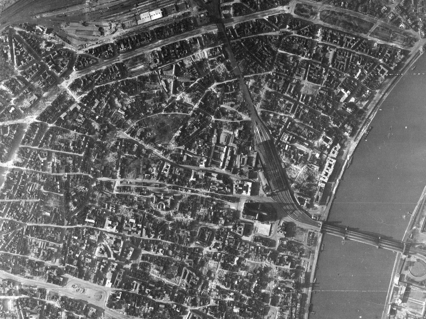 A black and white aerial photo of the city of Cologne, with a wide river crossed by a railway bridge on the right. The streets are visible, and some buildings have been damaged by bombing. Cologne cathedral is visible just below right of centre.