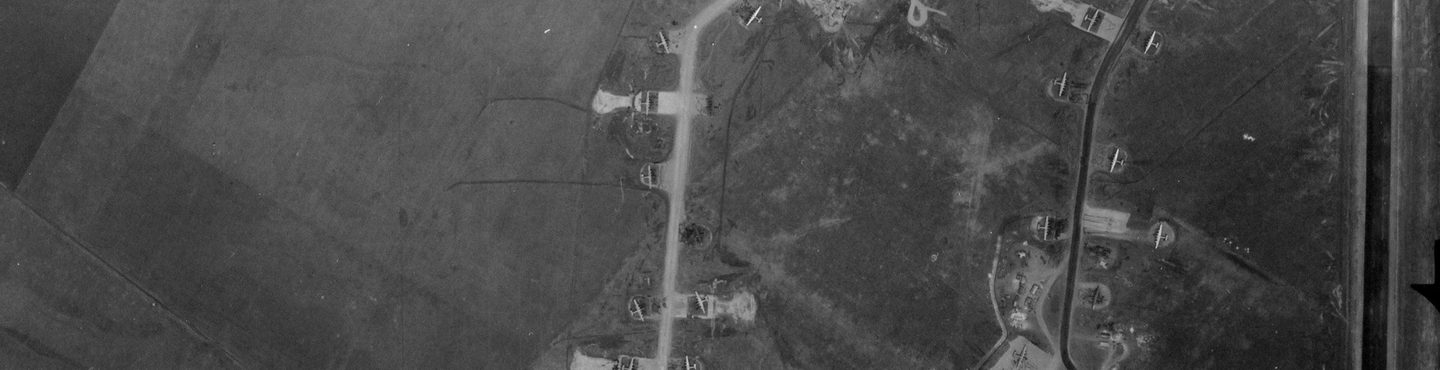 A black and white aerial photo of 17 planes at an airfield. The planes are all situated in bays off two tracks running vertically through the image – one light-coloured and one dark. The planes and tracks are surrounded by featureless fields.