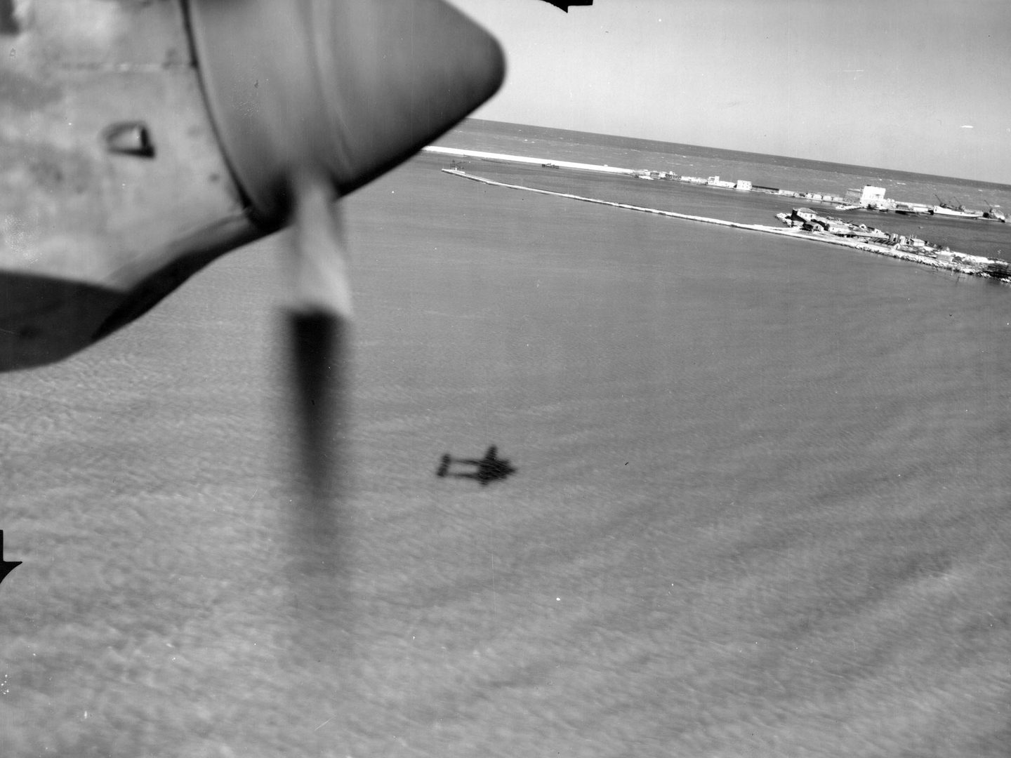 A black and white oblique aerial photo of the sea, with a strip of sky at the top. A harbour is in the distance and the shadow of the reconnaissance plane is visible on the sea. In the top left corner a portion of plane propeller can be seen.