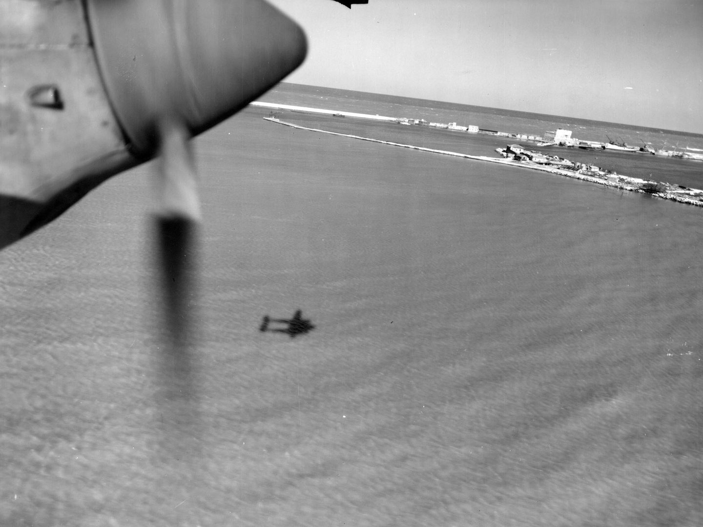 A black and white oblique aerial photo of the sea, with a strip of sky at the top. A harbour is in the distance and the shadow of the reconnaissance plane is visible on the sea. In the top left corner a portion of plane propeller can be seen.