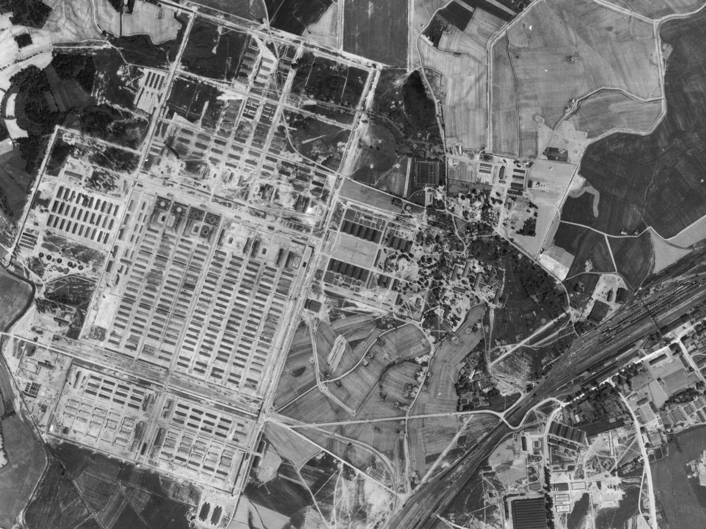 A black and white aerial photo of a large rectangular camp filled with numerous rows of huts. Smaller sets of huts sit to the left and right of the main camp. A railway line cuts across the bottom right corner. Fields are visible in the top right.