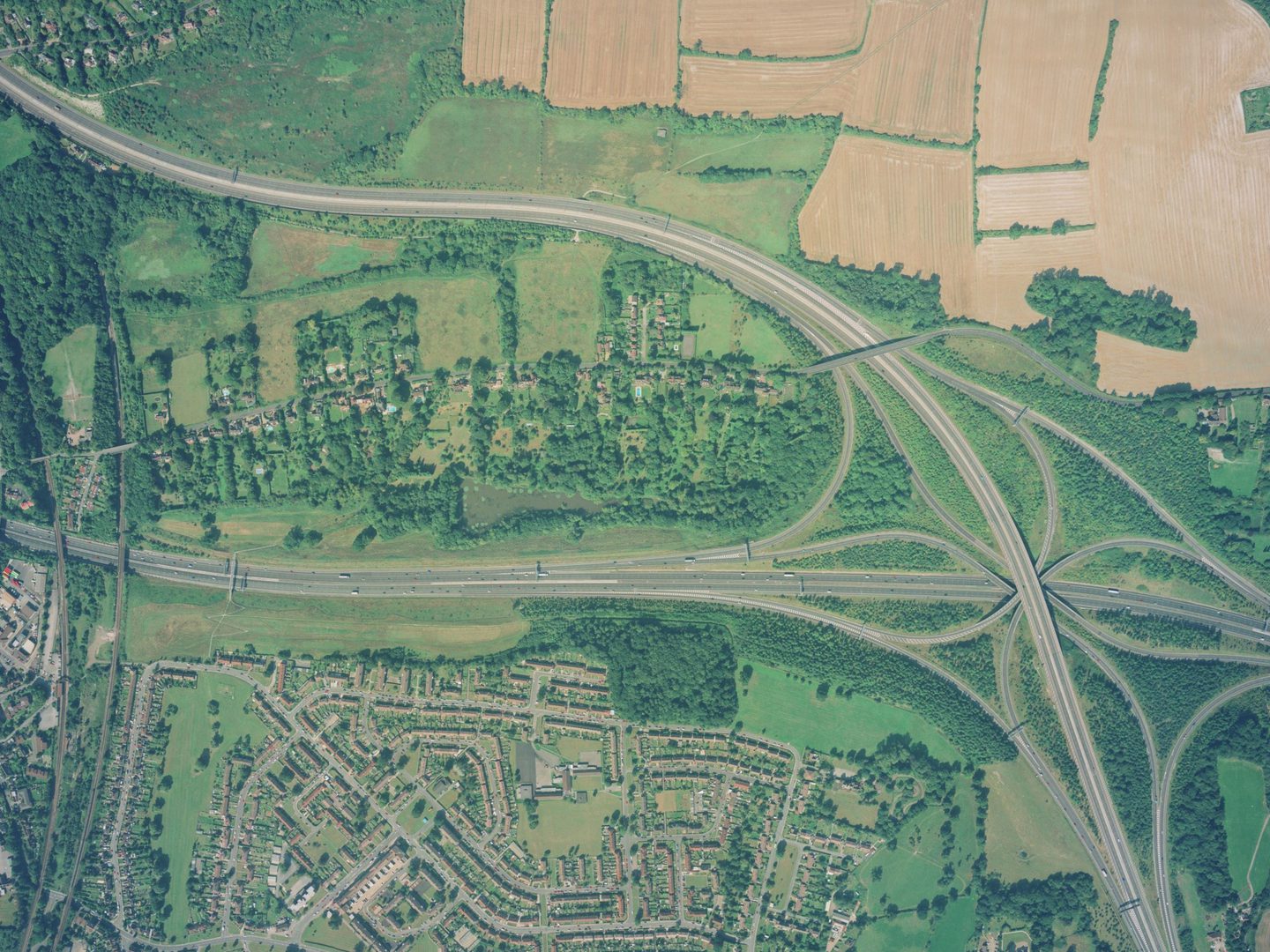 A colour aerial photo of a motorway junction. Brown fields occupy the top right corner, there are green trees in the middle and houses along the bottom edge. The junction is to the right, with two parallel roads leading away from it to the left edge.