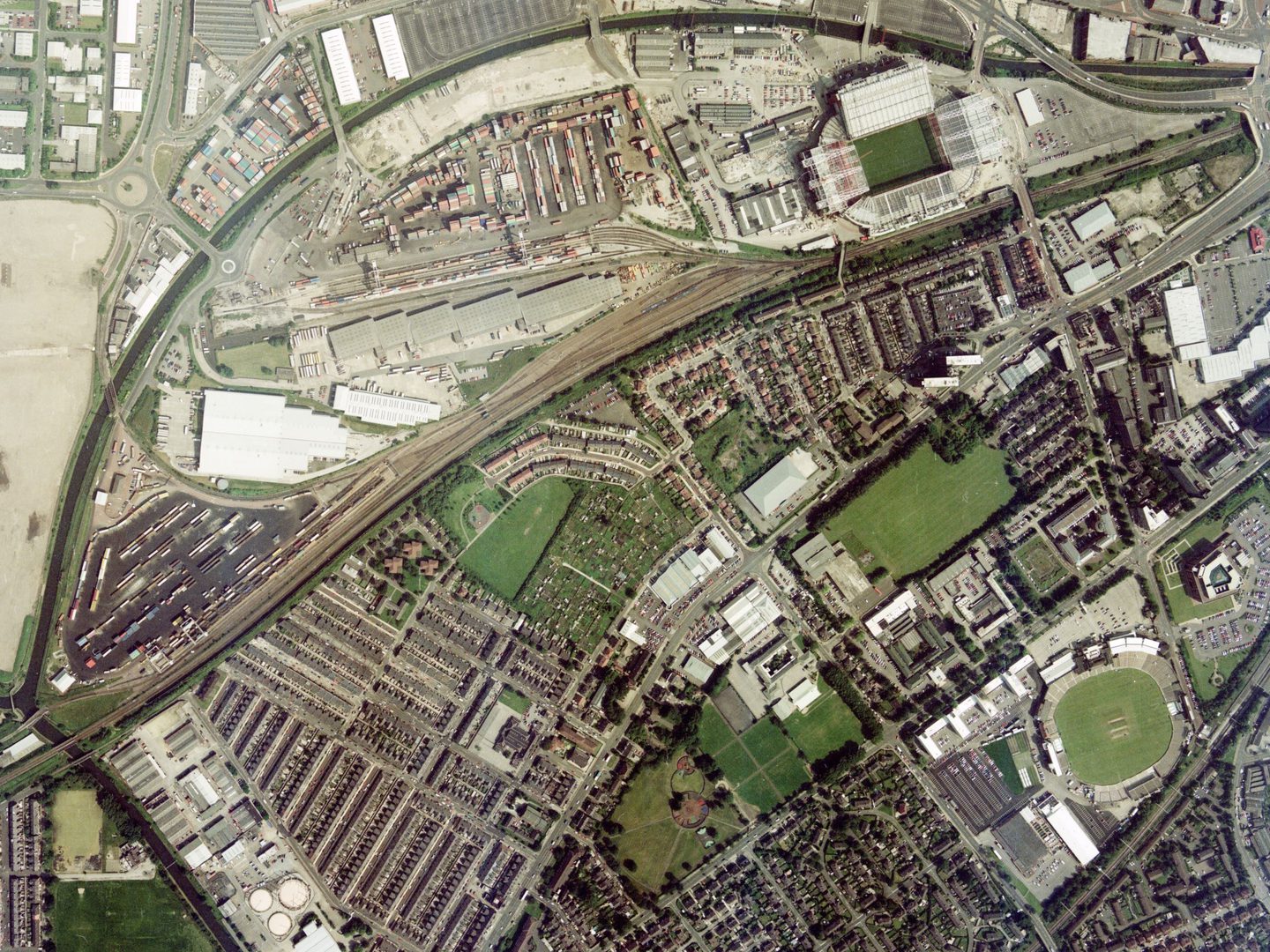 A colour aerial photo of an industrial part of Manchester, with a canal and railway running diagonally through the top half of the image. The football stadium is top right, and the cricket ground bottom right. They stand out as green rectangles.