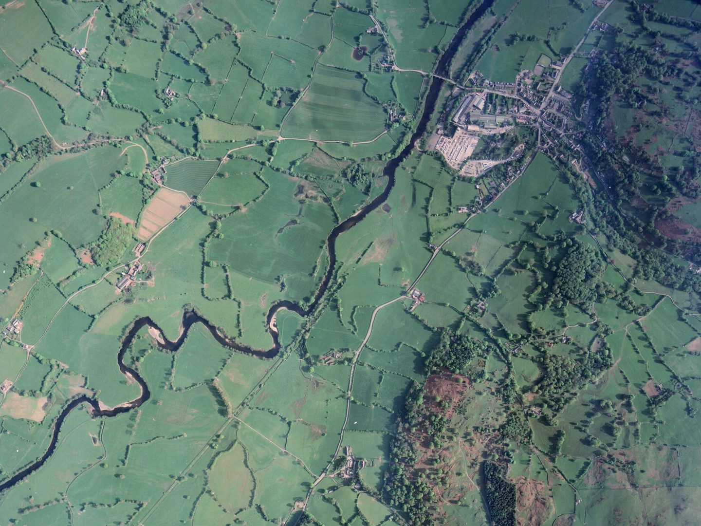 A colour aerial photo of countryside. A dark blue river meanders from the bottom left corner to the top right. There are wooded hills to the right and a small village in the top right corner. The green fields are divided by hedgerows and trees.