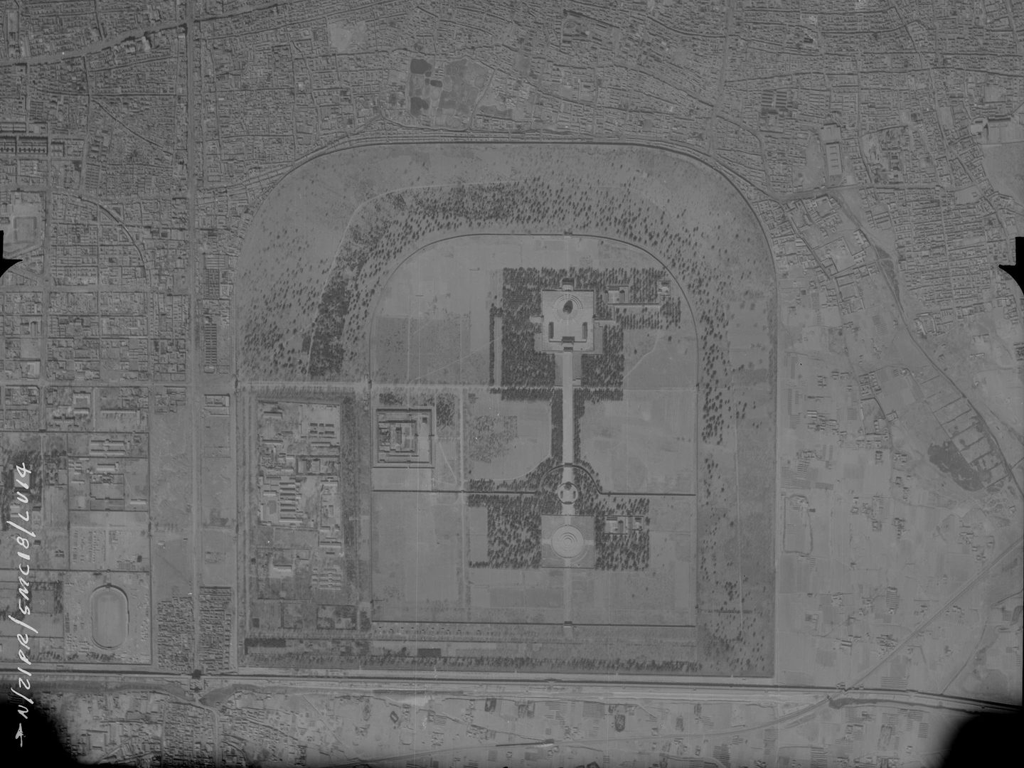 A black and white aerial photo of an expansive palace complex. The complex is a square shape with rounded top corners in the middle of the image. Densely packed buildings fill the top edge and top corners of the image. The image is quite pale.