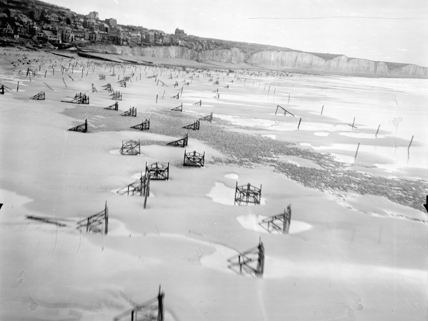 A low-level black and white oblique aerial photo of a beach. There is a town on some white cliffs in the background. The beach is scattered with numerous wooden structures to make landing more difficult. The sea is visible to the right of the image.