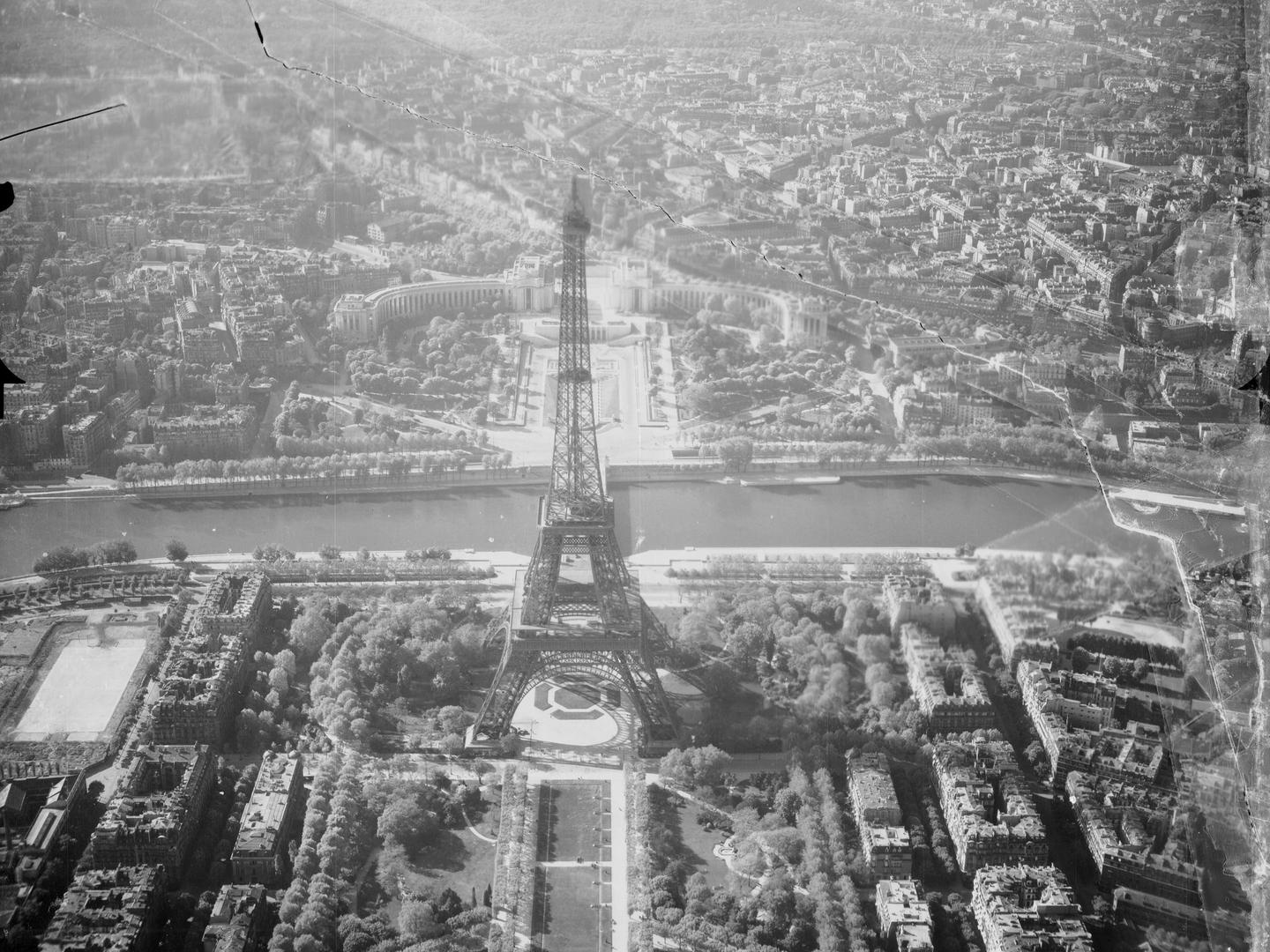 A black and white oblique aerial photo of Paris. The elegant metal lattice of the Eiffel Tower rises in the centre. A park with trees is in the foreground and a river runs horizontally behind the Eiffel Tower. Paris stretches into the background.