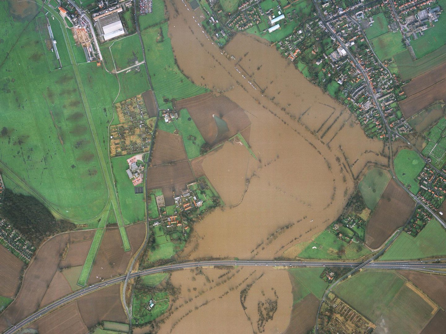 A colour aerial photo of a river running from top to bottom. The river has flooded and muddy water covers the fields either side. There are houses in the top right corner and a road runs across the bottom. There are green fields on the left side.