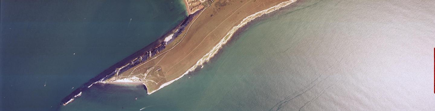 A colour aerial photo of the sea, very dark blue to the left and white from the glare of the sun on the right. In the middle a thin peninsula covered in brown grass extends into the sea. It has white cliffs and ends in a series of individual stacks.