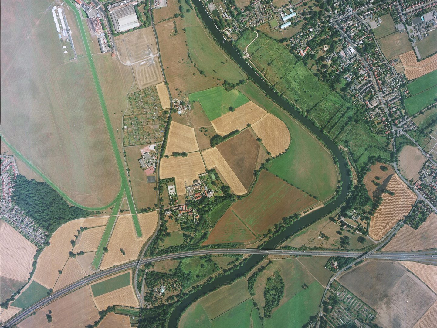 A colour aerial photo of a river running from top to bottom. The river has not flooded. There are houses in the top right corner and a road runs across the bottom. There are green and brown fields on the left side.