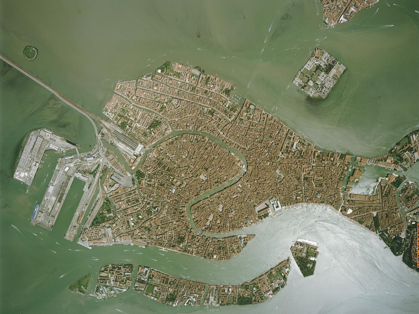 A colour aerial photo of Venice. Densely packed red roofs sit on an island surrounded by a lagoon of green-grey water. A railway joins the island from the image’s left edge. A large canal curls through the centre of the island.