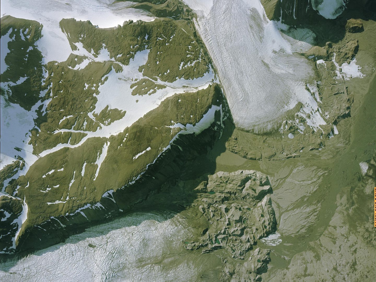 A colour aerial photo of a muddy brown landscape. Patches of white snow and ice are visible to the top left. At the top right and bottom left two white tongues of ice stretch into the brown landscape.