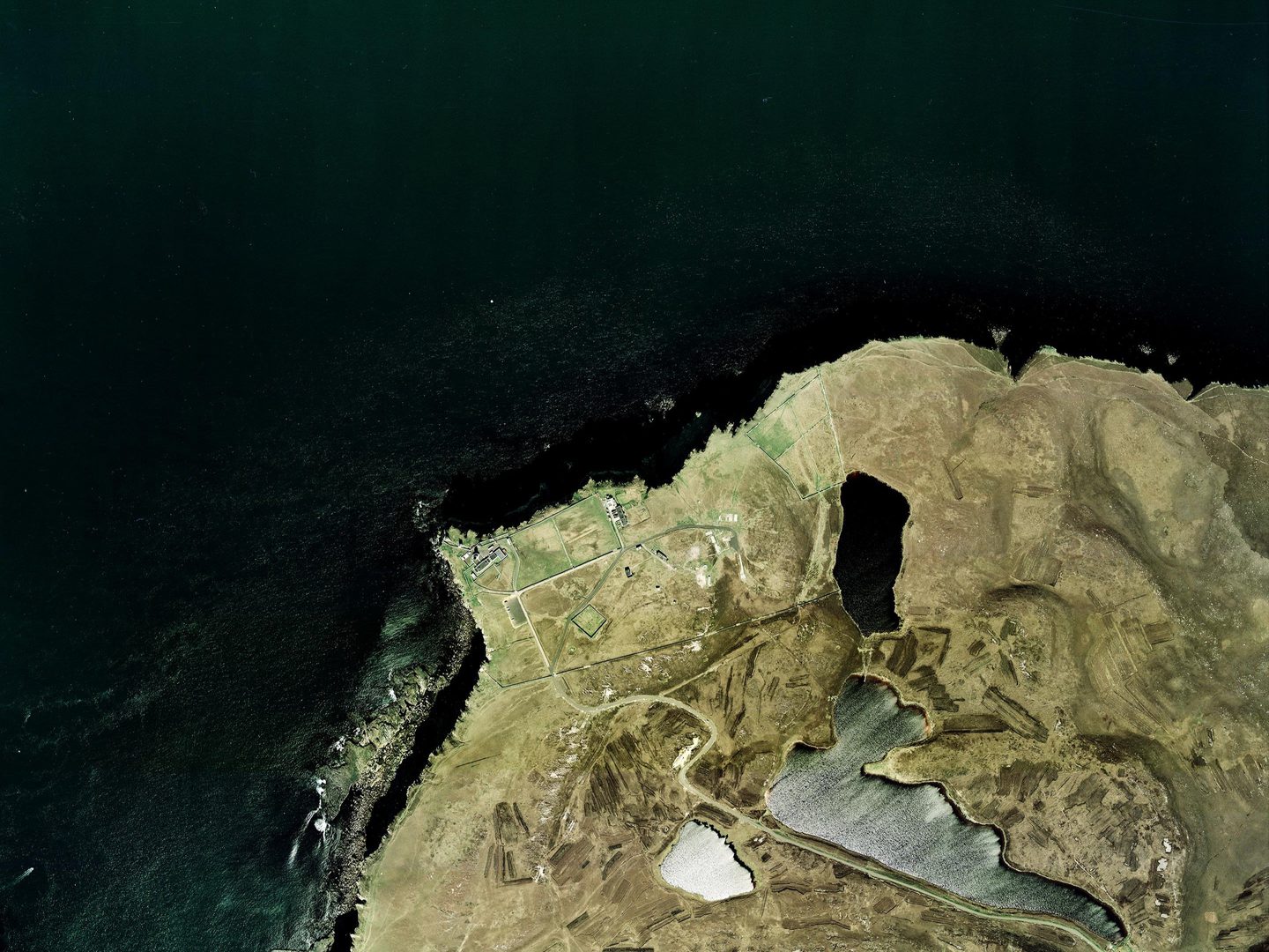 A colour photo of a grassy headland protruding into a very dark blue sea. The headland is semi-circular and covered in brown grass. There are three small lochs on the headland, and a small complex of buildings right at the water’s edge.