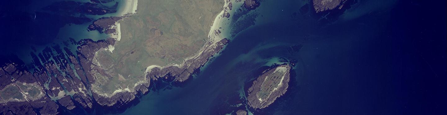 A colour aerial photo of islands surrounded by a deep blue sea. There is one big island left of centre and a small one to the right. The islands have green grass and a mix of rocky and sandy shores. The sea is light blue over the sand.