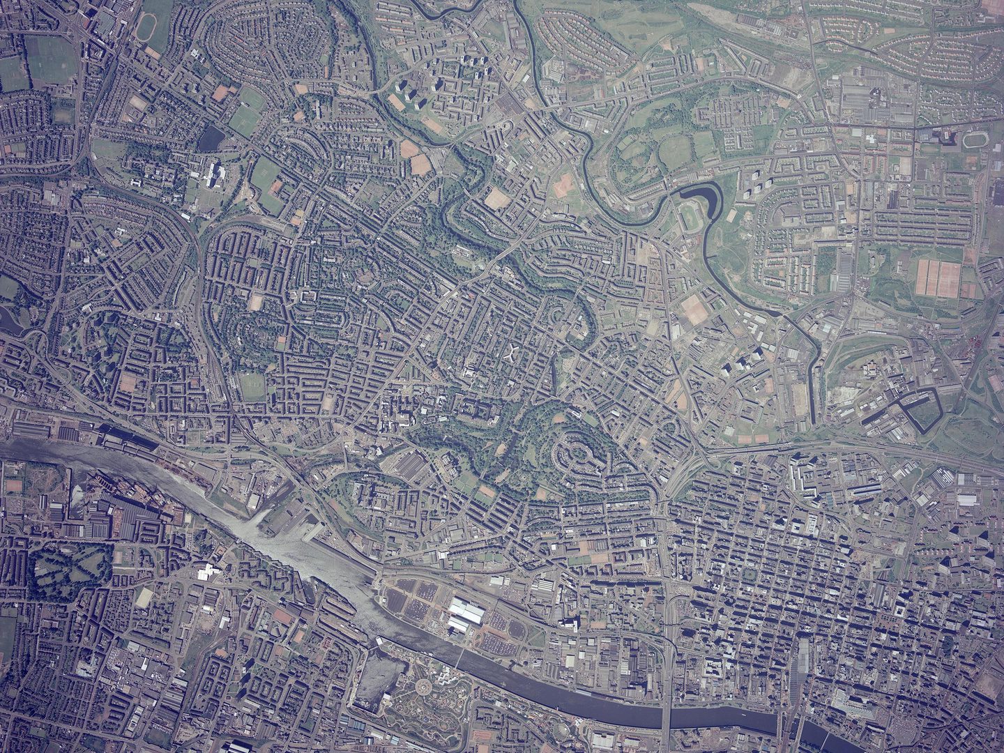A colour aerial photo of Glasgow from a high altitude. Grids of streets are visible across the image, with extensive green spaces in the top right corner. A large river runs along the bottom edge, with smaller rivers running vertically to meet it.