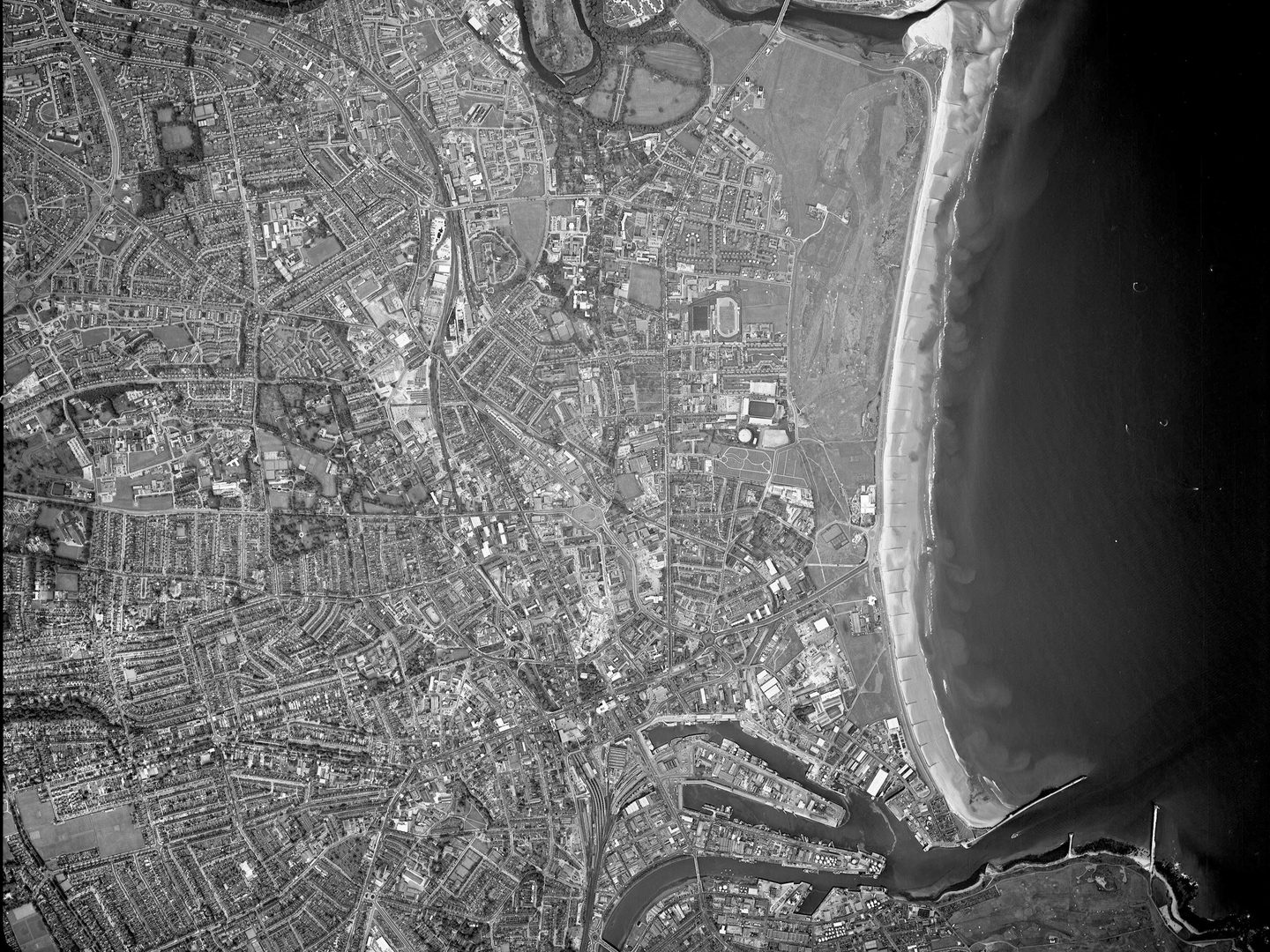 A black and white aerial photo of Aberdeen, with the city occupying the left-hand two thirds. The dark sea occupies the right third, with a pale beach between it and the city. The city is densely packed with buildings, with a river at the bottom.